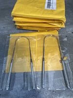 Two stainless steel tongue scrapers in clear plastic packaging, placed on yellow paper packaging, showing the front side with smooth handles and grooved ends.