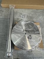 Photo showing the vertical rod of the stainless steel paper towel holder wrapped in plastic next to the boxed base and screws.