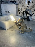 Mouse-shaped table lamp with large Edison bulb lit on a grey surface near a patterned wall socket.