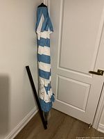 Blue and white striped folded umbrella leaning against a wall with black pole stand next to it.