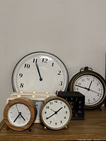 Shows 5 clocks including large round wall clock with white face, 2 small round clocks with wood and brass frames, black rectangular alarm clock, and cream flip clock.