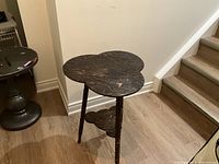 Full view of the clover-shaped table showing the top, legs, and lower shelf in a room corner with wooden floor and baseboard.