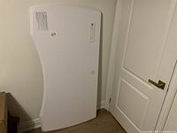 Image showing the front surface of the white ergonomic desk top, leaning against a wall near a white door. Curved contour edge visible.