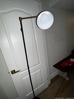 Full view of the tall industrial style floor lamp with black metal stand and white shade, angled upwards, positioned near a door.