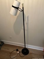 Full view of black metal floor lamp with white adjustable shade against a plain wall and wooden floor.