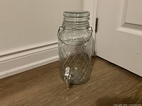 Front view of a clear glass beverage dispenser with diamond pattern, metal screw lid, and attached spigot placed on wooden floor near white baseboard and door.