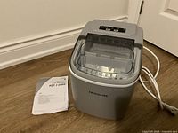 Front and top view showing the ice maker with transparent lid and control panel, alongside the instruction manual.