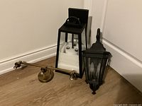 Three lighting fixtures on floor: black lantern, ornate black wall lantern, brass adjustable wall light