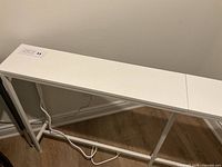 Side view of the long thin white desk showing the minimalist metal legs and the power cord running along the floor.