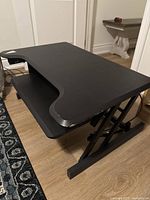 Side angled view showing the black adjustable desk riser with ergonomic curved desktop surface and sturdy metal frame on wooden floor.