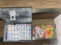 White Mah Jong tiles with traditional markings placed in a metal storage box with lid open. Next to the box is a plastic bag containing round game chips in multiple colors.