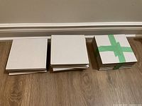 Three stacks of brand new medium square grey tiles placed on wooden floor against a baseboard wall.