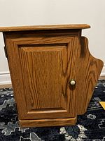 Close-up image of the oak end table showing the paneled cabinet door with brass knob and magazine rack side panel with curved cutout design.