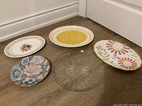 Five assorted plates arranged on floor showing diverse floral patterns and one clear glass plate