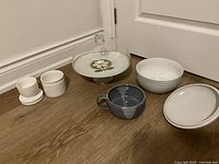 Photo showing all six items including two small white bowls with saucers, decorative floral plate with glass dome, white ridged bowl, bluish-gray mug, and white lidded bowl.