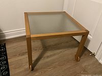 Square coffee table with light wooden frame and frosted glass top, showing the table surface and legs with wheels on one side.