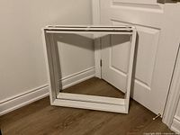 Two white steel rectangular table legs positioned standing upright against wall and door. Shows entire leg structure with mounting holes visible.