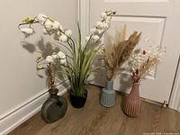 Four vases arranged on floor beside a white door. From left to right: greenish-gray rounded vase with dried brown stems, black pot vase with white orchid stems, light blue ribbed vase with pampas grass, pink ribbed vase with dried mixed flowers.