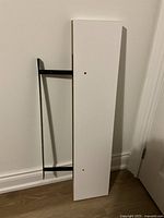 Photo showing the white rectangular shelf panel with black metal bracket leaning against wall, front view.