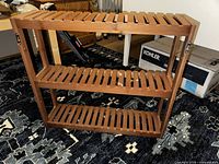 Front view of the wooden three-tier slatted shoe rack placed on a patterned carpet with surrounding items.