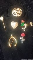 Seven assorted brooches displayed on a black velvet surface showing various designs including filigree, heart, rose, golf, and wishbone.