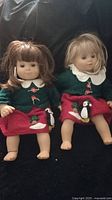Two American Girl twin dolls wearing matching green sweaters and red skirts with penguin decorations, shown against a black background.