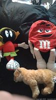Image showing three plush items: Marvin The Martian plush, red M&M plush backpack, and Ganz Webkinz cougar plush lying on a black surface.