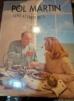 Cookbook titled 'Pol Martin: Love at First Bite' featuring man feeding woman at a table, glossy cover.