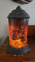 Front view of lit motion lamp showing detailed forest fire graphic on cylindrical glass with cast metal ornamental top and base.