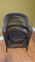 Front view of antique dark brown wicker chair showing open lattice weave on backrest, solid caned seat and curved armrests.