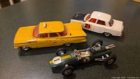 Lot of three Corgi die-cast cars: yellow Chevy Impala taxi, white Rover 2000 sedan, green Lotus Formula 1 race car with driver figure visible on dark surface.
