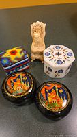 Five small decorative boxes arranged together: two black round Russian lacquer boxes with colorful artwork, a carved bone cat figurine box, a white hexagonal marble box with floral inlay, and a square beaded box with geometric pattern.