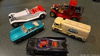 Group photo showing all five toy cars: Batmobile (black), green Ferrari, milk truck (cream), white 1928 Merc Benz, and red 1912 Packard Landaulet.