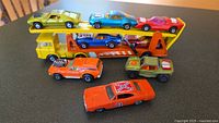Front view of the yellow car carrier truck loaded with eight colorful toy cars and one separate orange 'Dukes of Hazzard' Dodge Charger toy car in front.