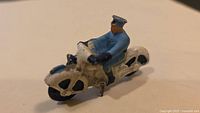 Side view of the metal policeman figure seated on a white motorcycle, painted blue uniform and black shoes visible, showing details and paint wear.