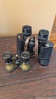 Two vintage binoculars on a wooden surface, larger black Birks 7x35 with leather strap and smaller metal Perfection Glass binoculars, side view.