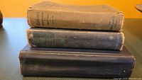 Stack of three vintage books showing condition of covers and spines. Top book has a tan cloth spine and cover, middle and bottom books have dark cloth covers.