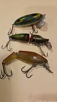 Photo showing the three vintage fishing lures, one green L&S Pikemaster and two unmarked smaller lures with jointed bodies and metal treble hooks.
