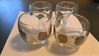 Four small vintage drinking glasses with gold and blue circular graphics arranged on black surface with white paper towels inside, showing overall lot contents and condition.