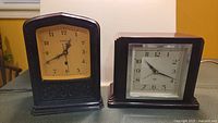 Two vintage bakelite electric table clocks side by side, Hammond on left with yellowed face and filigree below, Smith Sectric on right with square face and deco design
