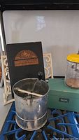 Photo showing the vintage metal flour sifter, wooden recipe box, and the hardcover recipe book on a decorative stand