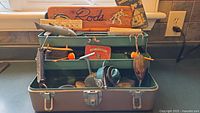 Photo showing open green metal Lucky Line tackle box with multiple vintage lures, fishing reel and bobbers inside, key visible on top compartment.