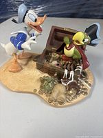 Front view showing Donald Duck reaching toward pirate parrot and open treasure chest on sand base