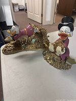 Two resin figurines on a white table—one Scrooge diving into coins, one standing holding coins.