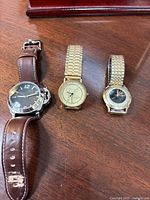 Three watches laid out on wood surface showing large silver-tone leather strap watch, gold-tone Wittnauer in center, smaller Hamilton on right