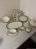 Full set of white porcelain dishes with gold trim displayed on table including plates, serving pieces, teapot, creamer, sauce boat, and stand.