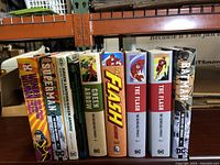 Six hardcover omnibuses on shelf showing Wonder Woman, Superman, Green Lantern & Green Arrow, two The Flash volumes, and Batman