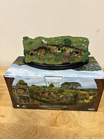 Front view of the Hobbit hole model with detailed moss-covered roof, round door, windows, and garden accents, displayed on its original box.