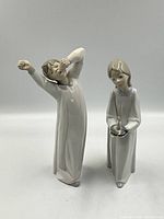 Front view of both Lladro porcelain figurines, girl holding candle and boy yawning in pajamas.