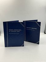 Two closed blue coin folders side by side with gold-stamped titles